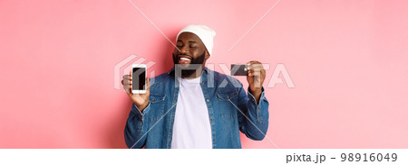 Online shopping. Satisfied Black man nod in approval, smiling and looking at phone, showing credit card and smartphone screen, standing over pink background 98916049