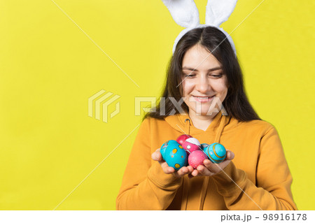 A happy brunette woman with rabbit ears on her head holds Easter eggs and smiles. Prepare for Easter, on a yellow background with copy space for text. A happy brunette woman with rabbit ears on her head holds Easter eggs and smiles. Prepare for Easter, on a yellow background with copy space for text. 98916178
