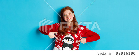 Winter holidays and Christmas Eve concept. Pretty redhead girl pointing fingers at cute xmas sweater with penguin, standing over blue background 98916190