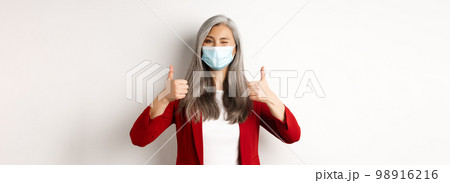 Coronavirus and business concept. Asian female entrepreneur in face mask looking cheerful, showing thumb-up in approval, white background Coronavirus and business concept. Asian female entrepreneur in face mask looking cheerful, showing thumb-up in approval, white background 98916216