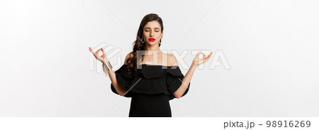 Calm and patient young brunette female meditating, standing in black dress with hands in zen gesture, standing against white background 98916269