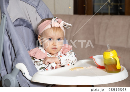 A beautiful one-year-old girl eats cookies sitting in a feeding chair. Complementary foods for children, sweets, sugar. A beautiful one-year-old girl eats cookies sitting in a feeding chair. Complementary foods for children, sweets, sugar. 98916343