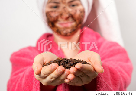 A woman in a pink bathrobe with a coffee mask on her face holds coffee beans in her hands 98916548