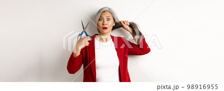Asian senior woman holding scissors and looking nervous at camera, thinking about cutting hair, changing hairstyle, standing over white background 98916955