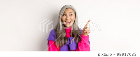 Close-up of stylish korean senior woman with grey hair looking and pointing upper left corner, showing promotion offer, standing over white background 98917035