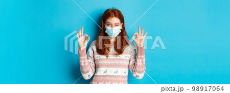 Winter, coronavirus and social distancing concept. Cheerful redhead girl in face mask, showing okay signs and winking, recommending excellent product, blue background 98917064