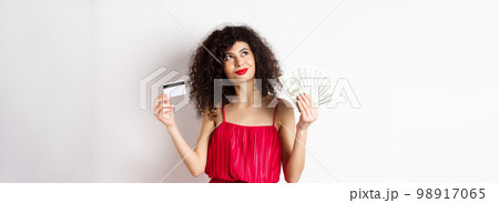 Shopping. Woman thinking and smiling, holding money with plastic credit card, wearing red elegant dress and evening makeup, white background Shopping. Woman thinking and smiling, holding money with plastic credit card, wearing red elegant dress and evening makeup, white background 98917065