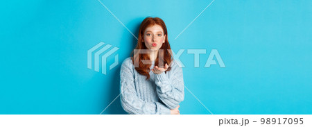 Lovely teen girl in sweater blowing air kiss, pucker lips and staring at camera, standing against blue background 98917095