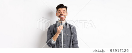 Handsome positive guy showing white perfect smile with magnifying glass, standing against white background Handsome positive guy showing white perfect smile with magnifying glass, standing against white background 98917126