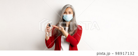 Covid, pandemic and business concept. Asian female manager in medical mask using hand sanitizer and smiling at camera, standing with antiseptic over white background 98917132