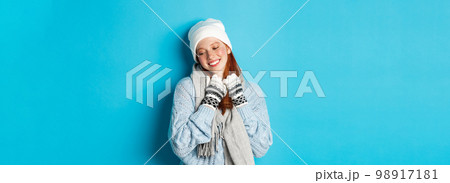 Winter and holidays concept. Cute redhead girl blushing and looking away dreamy, wearing warm beanie, sweater and scarf with gloves, standing over blue background 98917181