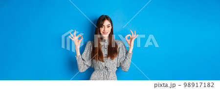 Confident adult woman in dress with natural hair color, showing okay signs and smiling, like and approve something good, standing on blue background Confident adult woman in dress with natural hair color, showing okay signs and smiling, like and approve something good, standing on blue background 98917182