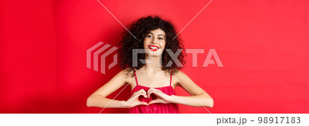 Lovers day. Beautiful woman celebrating valentines, showing heart sign and smiling, standing in romantic red dress on studio background 98917183