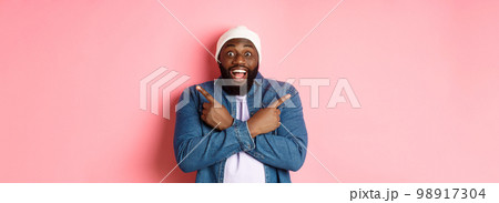 Happy young african-american hipster guy pointing fingers sideways, smiling and showing two choices, showing offers, standing over pink background 98917304