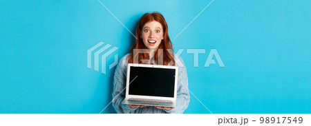 Excited redhead female freelancer showing laptop screen, staring at camera amazed, standing with computer against blue background Excited redhead female freelancer showing laptop screen, staring at camera amazed, standing with computer against blue background 98917549