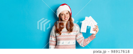 Holiday promos and real estate concept. Cheerful redhead woman in santa hat holding paper house in hand and smiling, standing in sweater against blue background Holiday promos and real estate concept. Cheerful redhead woman in santa hat holding paper house in hand and smiling, standing in sweater against blue background 98917618