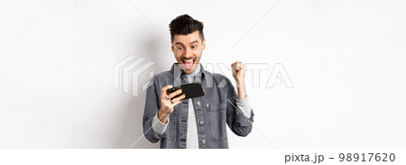 Happy man shouting yes, winning video game on mobile phone and triumphing, achieve online goal, standing on white background Happy man shouting yes, winning video game on mobile phone and triumphing, achieve online goal, standing on white background 98917620