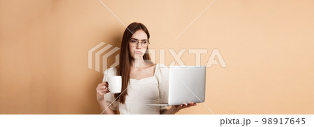 Tired working girl looking at laptop bored, drinking coffee while using computer for work, standing on beige background Tired working girl looking at laptop bored, drinking coffee while using computer for work, standing on beige background 98917645