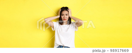Asian woman holding hands on head and looking sad, having a problem, standing anxious against yellow background 98917669