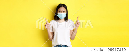Covid-19, pandemic and social distancing concept. Disappointed asian girl in medical mask, frowning upset and pointing fingers up at logo, standing over yellow background Covid-19, pandemic and social distancing concept. Disappointed asian girl in medical mask, frowning upset and pointing fingers up at logo, standing over yellow background 98917830