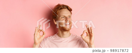 Close up of handsome redhead man in t-shirt, smiling satisfied and looking left, showing okay signs in approval, praising awesome product, standing over pink background Close up of handsome redhead man in t-shirt, smiling satisfied and looking left, showing okay signs in approval, praising awesome product, standing over pink background 98917872