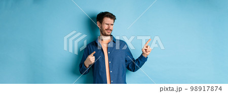 Skeptical young man pointing and looking left with disappointment, cringe from something bad, standing upset on blue background 98917874