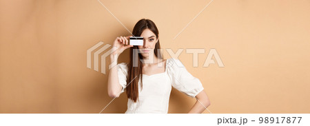 Sassy young woman showing plastic credit card on face and looking determined, going on shopping, standing against beige background Sassy young woman showing plastic credit card on face and looking determined, going on shopping, standing against beige background 98917877