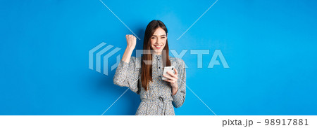 Happy pretty girl winning on mobile phone, smiling and chanting, holding smartphone, standing in dress against blue background 98917881