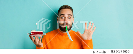 Close-up of funny adult man celebrating his birthday, holding bday cake with candle, blowing party wistle and rejoicing, standing over light blue background Close-up of funny adult man celebrating his birthday, holding bday cake with candle, blowing party wistle and rejoicing, standing over light blue background 98917906