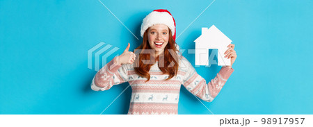Holiday promos and real estate concept. Satisfied female model with red wavy hair, wearing santa hat and sweater, showing paper house model and thumbs-up, blue background Holiday promos and real estate concept. Satisfied female model with red wavy hair, wearing santa hat and sweater, showing paper house model and thumbs-up, blue background 98917957
