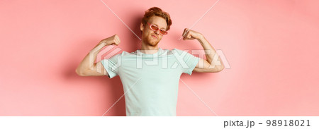 Confident young man with red hair, wearing summer sunglasses and t-shirt, showing strong and fit body muscles, flex biceps and staring cool at camera, pink background Confident young man with red hair, wearing summer sunglasses and t-shirt, showing strong and fit body muscles, flex biceps and staring cool at camera, pink background 98918021