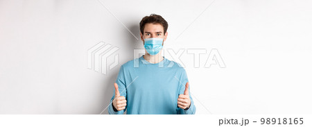 Covid-19, pandemic and social distancing concept. Young man in casual clothes and medical mask, showing thumbs up and looking serious, protecting from virus during quarantine Covid-19, pandemic and social distancing concept. Young man in casual clothes and medical mask, showing thumbs up and looking serious, protecting from virus during quarantine 98918165