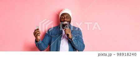 Handsome african-american man singing karaoke, reading lyrics on smartphone app and holding microphone, standing over pink background 98918240