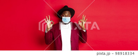 Covid-19 and fashion concept. Stylish african american man in hat and blazer, wearing face mask and showing okay sign, standing over red background Covid-19 and fashion concept. Stylish african american man in hat and blazer, wearing face mask and showing okay sign, standing over red background 98918334