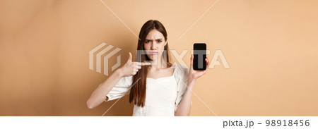 Disappointed frowning girl pointing at empty phone screen, complaining at online news, standing on beige background 98918456