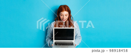 Amazed redhead girl staring at laptop screen and looking impressed, showing computer display, standing over blue background 98918458