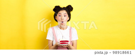Happy asian birthday girl with bright makeup, blowing candle on cake, making wish, standing on yellow background Happy asian birthday girl with bright makeup, blowing candle on cake, making wish, standing on yellow background 98918661