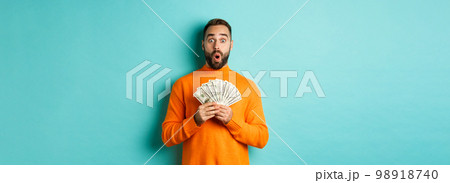 Photo of surprised guy holding money, looking amazed, standing with dollars against turquoise background Photo of surprised guy holding money, looking amazed, standing with dollars against turquoise background 98918740