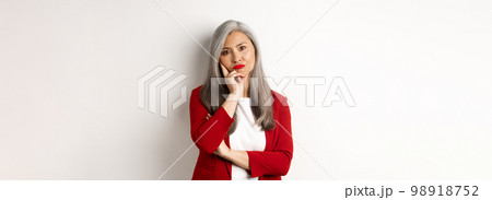 Annoyed and bothered asian businesswoman in red blazer, pouting and looking irritated at camera, standing over white background 98918752