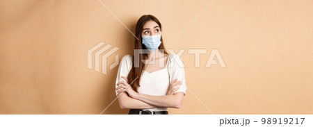 Pandemic and healthcare concept. Thoughtful girl in face mask looking at empty space logo, standing curious on beige background 98919217