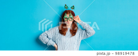 Winter holidays and Christmas sales concept. Beautiful redhead female model celebrating New Year, wearing funny party headband and glasses, smiling silly, blue background Winter holidays and Christmas sales concept. Beautiful redhead female model celebrating New Year, wearing funny party headband and glasses, smiling silly, blue background 98919478