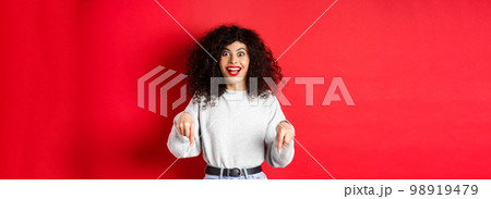 Excited woman pointing fingers down and smiling amazed, look here gesture, showing logo on red empty space Excited woman pointing fingers down and smiling amazed, look here gesture, showing logo on red empty space 98919479