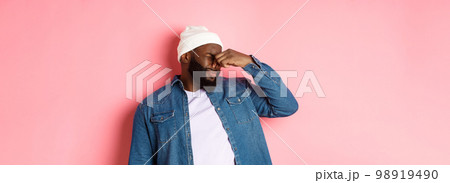Disgusted african-american man shut nose from aversion, displeased by awful bad smell, standing in beanie and denim shirt over pink background Disgusted african-american man shut nose from aversion, displeased by awful bad smell, standing in beanie and denim shirt over pink background 98919490