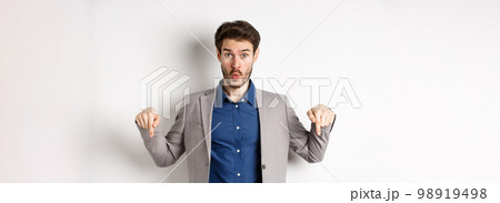 Wow look down. Excited business guy in suit pointing fingers bottom advertisement and look amazed, standing on white background 98919498