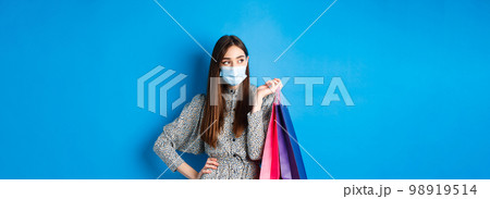 Covid-19, pandemic and lifestyle concept. Girl wearing medical mask on shopping in malls, holding paper bags and look aside at empty space, standing on blue background 98919514