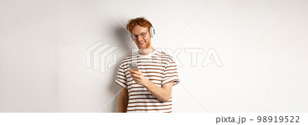 Technology concept. Young redhead hipster in glasses, listening music in headphoes, reading message on smartphone, white background Technology concept. Young redhead hipster in glasses, listening music in headphoes, reading message on smartphone, white background 98919522