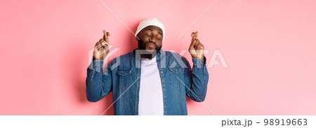 Hopeful and dreamy Black man making wish, cross fingers and smiling, standing over pink background 98919663
