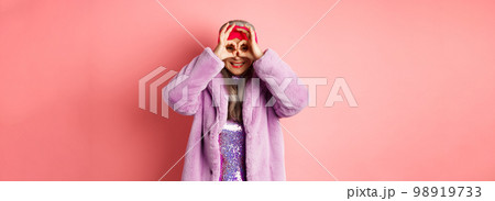 Fashion and shopping. Happy asian grandmother in stylish faur fur coat, looking through hand glasses, making binoculars and smilinga t camera, standing over pink background Fashion and shopping. Happy asian grandmother in stylish faur fur coat, looking through hand glasses, making binoculars and smilinga t camera, standing over pink background 98919733