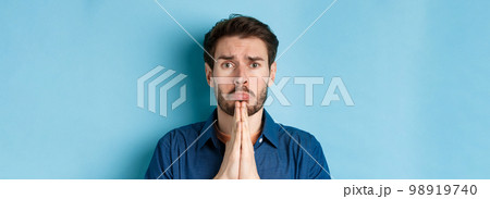 Sad young man begging for help, apoligizing and sobbing miserable, standing on blue background Sad young man begging for help, apoligizing and sobbing miserable, standing on blue background 98919740
