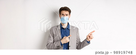 Covid-19, pandemic and business concept. Upset man in medical mask and office suit, frowning sad and pointing left at banner, standing on white background 98919748
